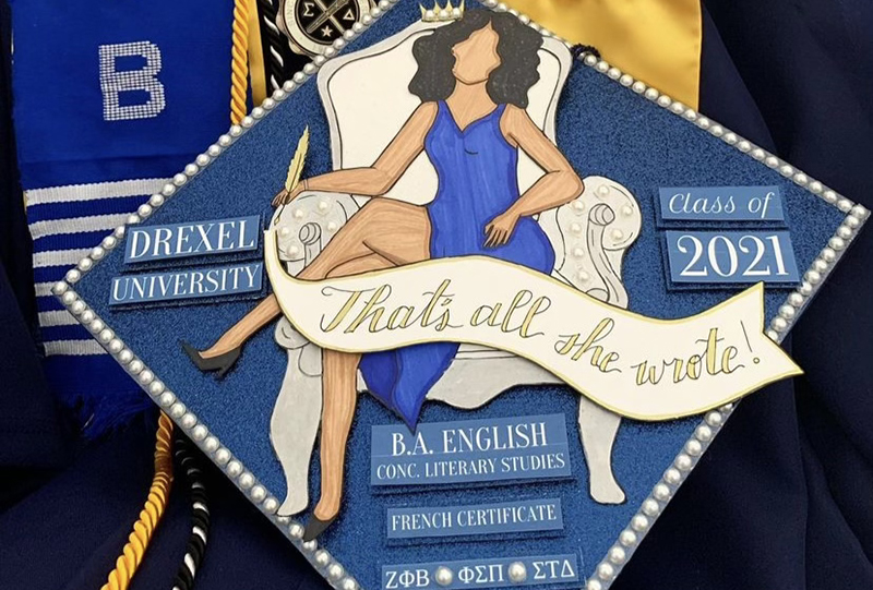 A commencement cap with the phrase "That's All She Wrote" and a seated woman on a chair.
